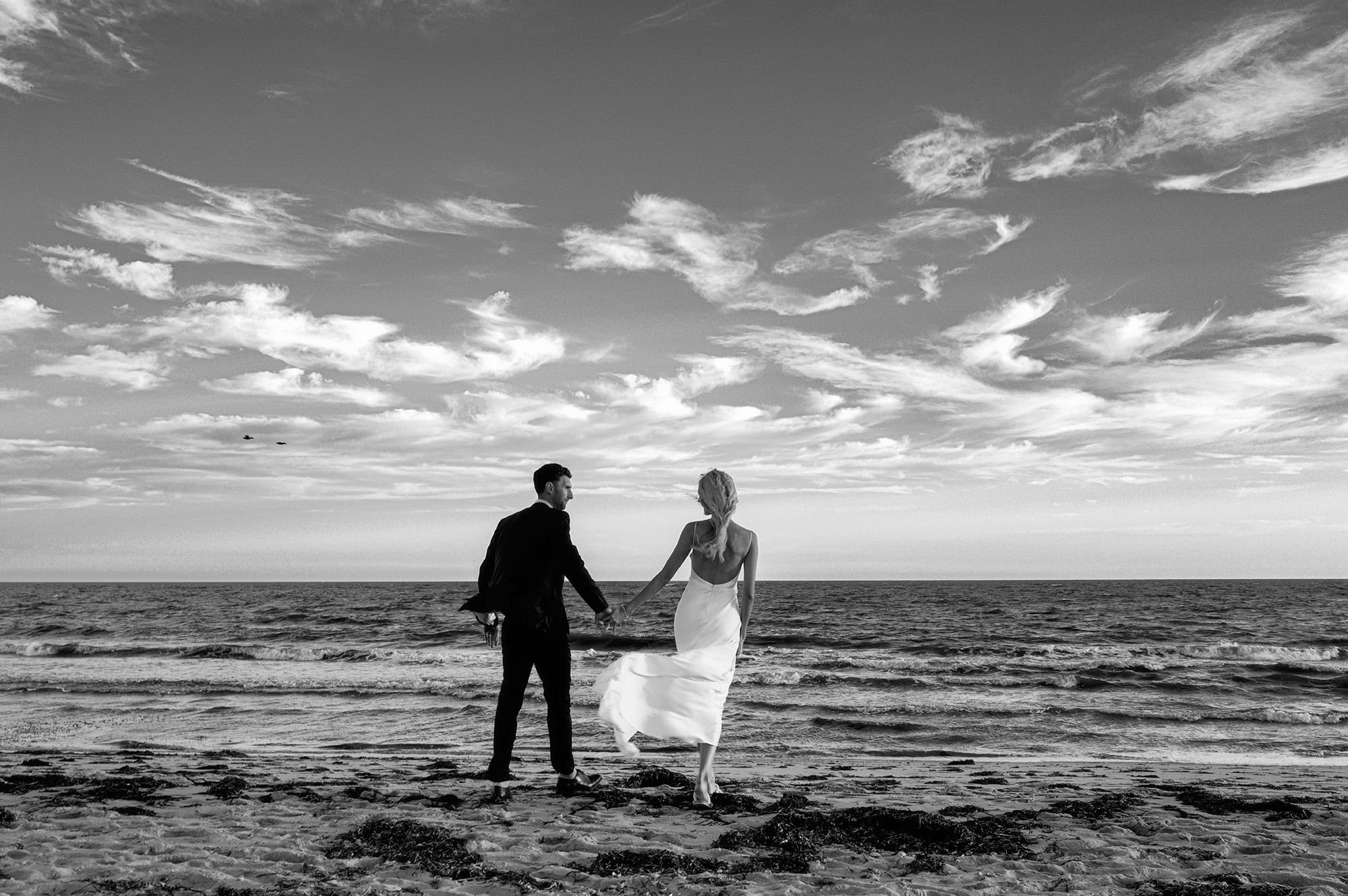 black and white photo of a caucasian groom and bride on the beach facing the water
